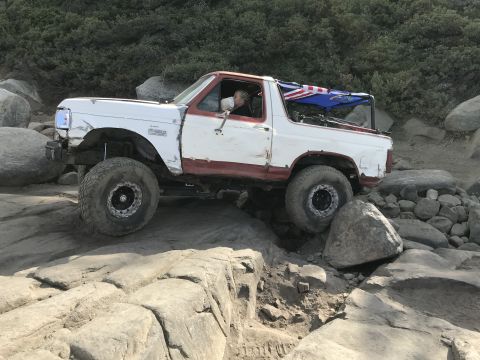 1984 Ford Bronco - A white and red Ford Bronco is navigating a rocky, off-road trail. The vehicle has large, knobby tires and a custom roll cage with flags at…