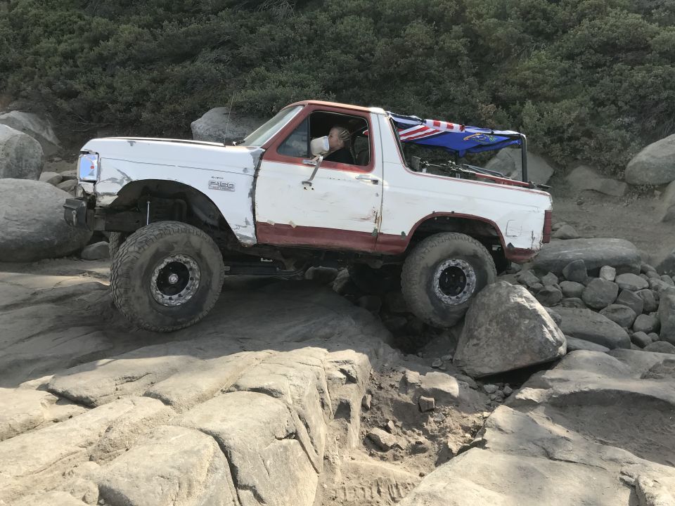White and red Ford Bronco off-roading on a rocky trail with large tires and a roll cage.