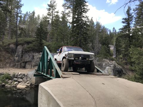 1984 Ford Bronco - A modified white Ford pickup truck with a red roof is driving across a narrow metal bridge over a body of water. The truck has large off-ro…