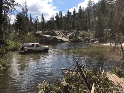 1984 Ford Bronco - A white Ford Bronco is driving through a shallow river in a wooded, rocky area. The vehicle appears to be an off-road build with modificati…
