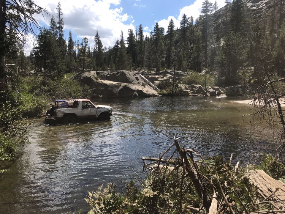 White Ford Bronco driving through a river in a forest.