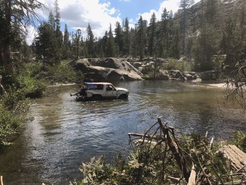 1984 Ford Bronco - A white and red 1984 Ford Bronco is driving through a shallow river surrounded by trees and rocks. Several people are riding in the bed of…