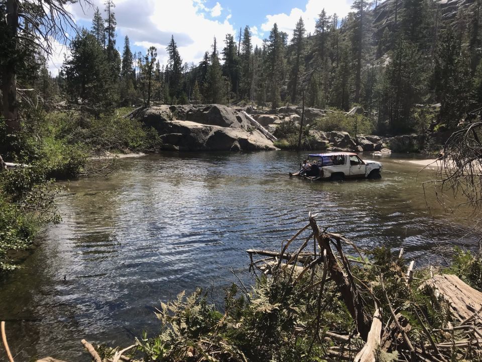 White 1980s Ford Bronco submerged in a shallow river, surrounded by trees and rocks.