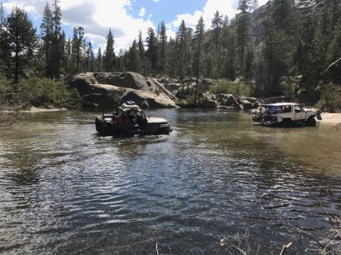 1984 Ford Bronco - Two off-road vehicles are crossing a shallow river in a wooded, rocky area. The vehicle in the foreground is a dark-colored Jeep, and the v…