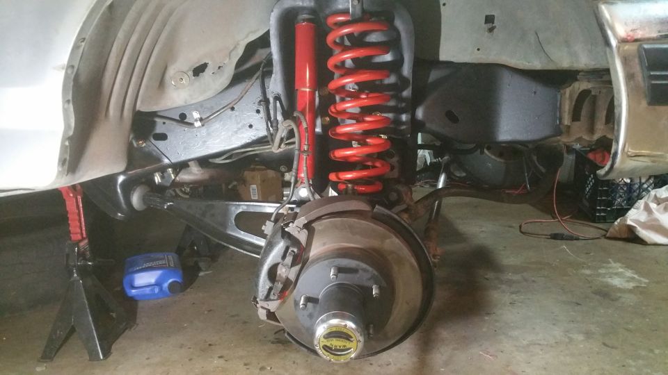 Front suspension of a vehicle with red coil spring and shock absorber, jack stand, and blue fluid jug.