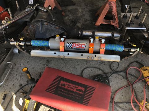 1992 Jeep Wrangler - A close-up view of a vehicle's front axle assembly, featuring a hydraulic steering cylinder with PSC branding, leaf springs, and various to…