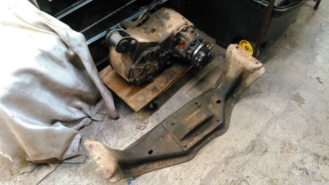 1978 Ford Bronco - A dirty transfer case sits on a wooden dolly next to a rusty metal crossmember. A light-colored cloth is draped over a part of the transfer…