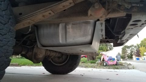1985 Ford Bronco - Underside view of a vehicle's fuel tank and rear axle, with a large metal skid plate protecting the tank. A pink toy tricycle is visible in…