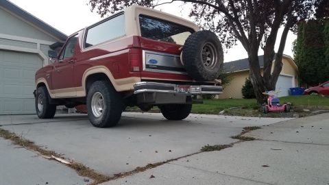 1985 Ford Bronco - The rear passenger side of a red and tan two-door Ford Bronco is parked on a concrete driveway in front of a garage. A spare tire is mounte…