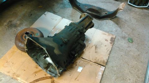 1978 Ford Bronco - A dark, greasy automatic transmission and torque converter sit on top of cardboard on a concrete floor. A black metal subframe is visible i…