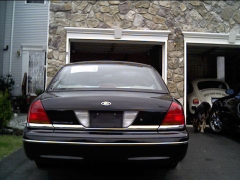 1999 Ford Crown Victoria - The rear of a black Ford sedan is shown parked in front of a garage, with other vehicles visible inside.