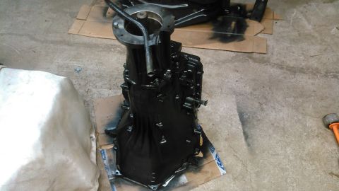 1978 Ford Bronco - A black painted transmission is shown resting on cardboard on a concrete floor. A white tarp is visible to the left of the transmission.