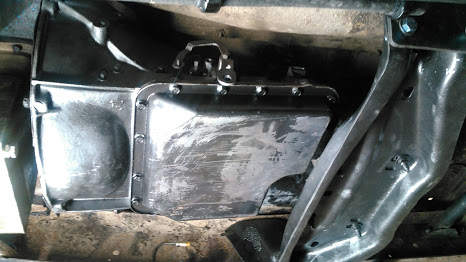 1978 Ford Bronco - The underside of a vehicle shows a black transmission pan with a silver bracket attached to the top. The pan is bolted to the transmission…