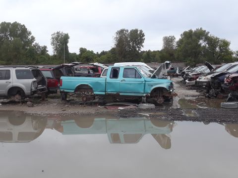 1994 Ford Bronco - A turquoise Ford F-150 pickup truck is parked in a junkyard with its hood open and missing wheels. Other vehicles and debris are visible in…