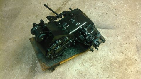 1978 Ford Bronco - A black, greasy transfer case is mounted on a wooden pallet, resting on a concrete floor. The transfer case has a lever protruding from its…
