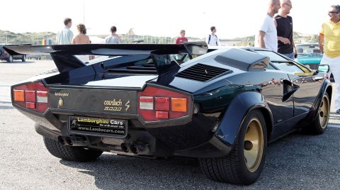 1992 Ford Bronco - Rear 3/4 view of a black Lamborghini Countach 5000 with a large rear wing and gold wheels. The car is parked outdoors on asphalt with peopl…