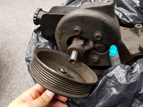 1978 Ford Bronco - A person is holding a ribbed pulley in front of a black power steering pump assembly. The parts appear to be dusty and are resting inside a…