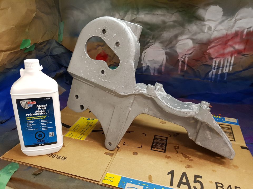 Bottle of metal prep next to a silver metal part, with a spray-painted drop cloth in the background.