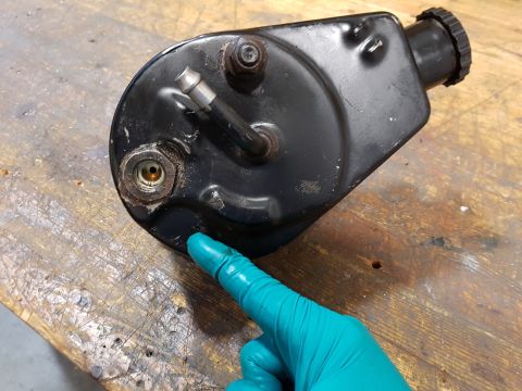 1978 Ford Bronco - A gloved hand points to a black power steering pump reservoir with a metal fitting and a black cap. The part is resting on a wooden surface.