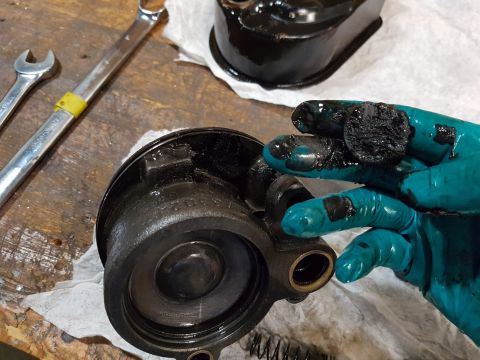 1978 Ford Bronco - A gloved hand holds a black, greasy component that appears to be a part of a vehicle's engine or braking system, with tools and other parts…