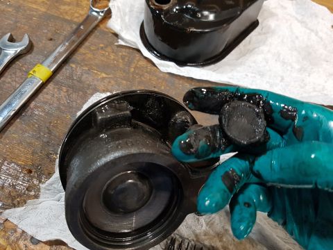 1978 Ford Bronco - A gloved hand holds a small, greasy, black circular part over a disassembled black metal component on a wooden workbench. Tools and paper t…
