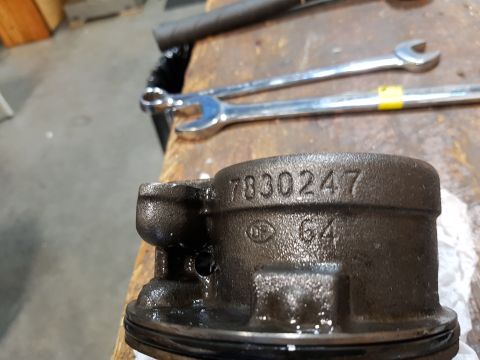 1978 Ford Bronco - A close-up of a dark, oily metal part, possibly a piston, with the numbers "7830247" and "64" stamped on its surface. The part rests on a w…