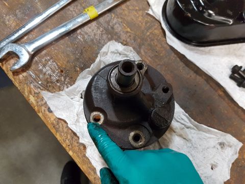 1978 Ford Bronco - A gloved hand points to a bolt hole on a black metal automotive part, likely a pump housing, resting on a wooden workbench next to a pair o…