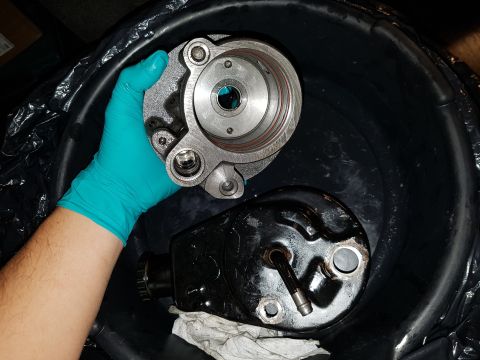 1978 Ford Bronco - A hand wearing a teal glove holds a silver metal part over a black plastic basin containing a black metal part and a white rag.