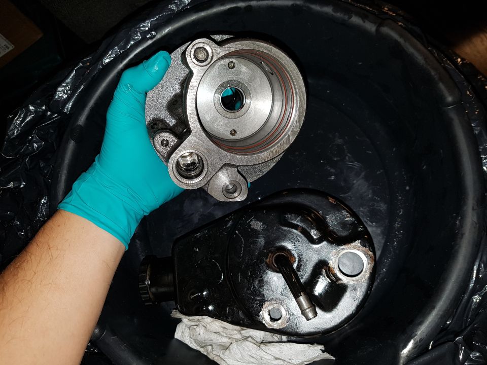 Hand in teal glove holding a silver metal part over a black basin with a black part and rag.