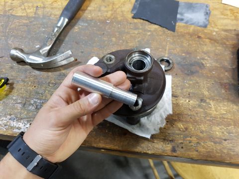 1978 Ford Bronco - A person's hand holds a metal cylindrical part over a dark-colored vehicle component on a wooden workbench. A hammer and sandpaper are visi…
