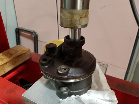 1978 Ford Bronco - A dark-colored automotive part, possibly a power steering pump housing, is being pressed into place by a metal cylinder. The part rests on…