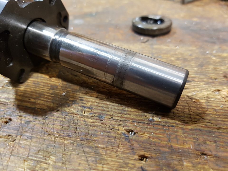 Metal shaft with wear marks and a dark band, on a wooden surface. Flanged component attached.