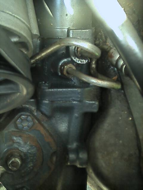 1989 Ford Bronco - Close-up looking down at a recirculating-ball steering gearbox mounted to the frame rail, with two curved metal hydraulic pressure and retu…