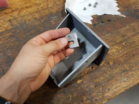1978 Ford Bronco - A hand holds a small metal bracket over a gray metal box on a wooden workbench. Several small ball bearings are visible on a piece of white…