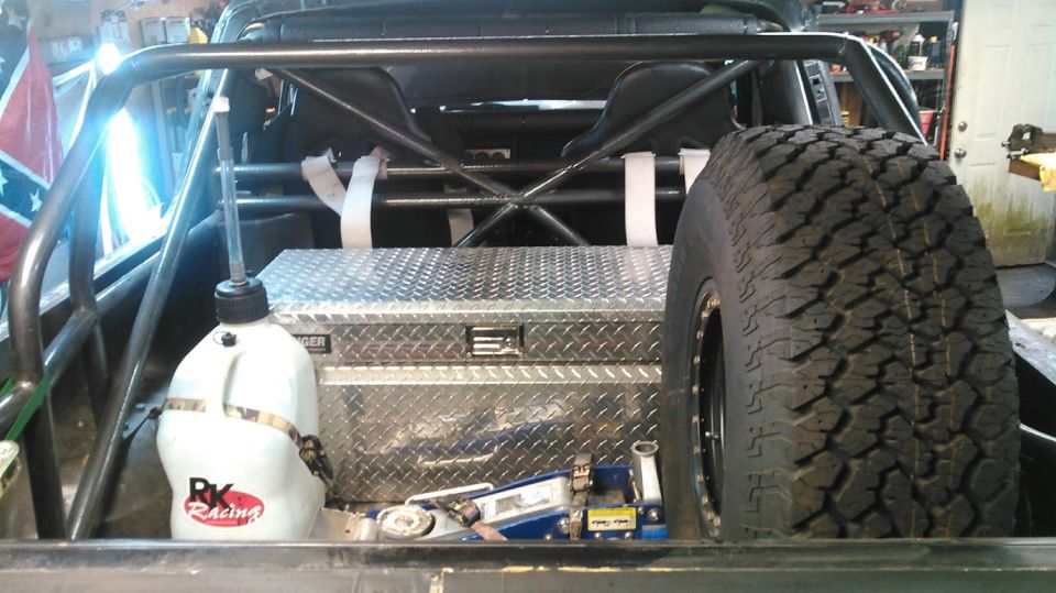 Interior of a modified vehicle with roll cage, racing seats, toolbox, spare tire, fuel jug, and jack.