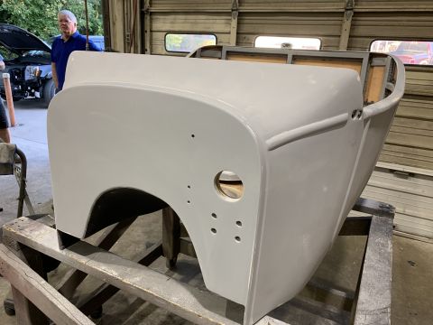 1923 Ford Model T - A light gray, unfinished automotive body, possibly a car or truck bed, sits on a wooden stand in a garage. A man in a blue polo shirt stand…