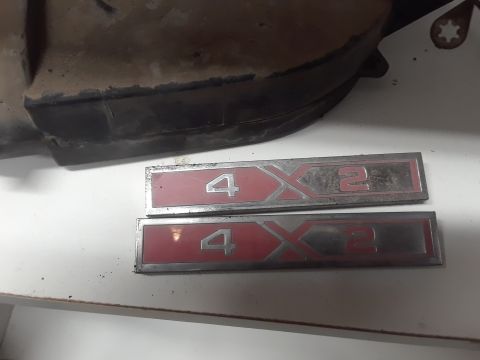 1994 Ford Bronco - Two identical chrome and red emblems that read "4X2" are shown on a white surface. A dark, dusty vehicle part is visible in the upper left…