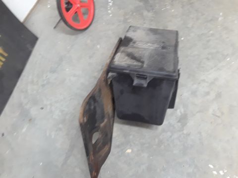 1994 Ford Bronco - A black plastic Ford-branded fuse box is mounted to a rusty metal bracket on a concrete floor. A red wheel is visible in the upper left cor…