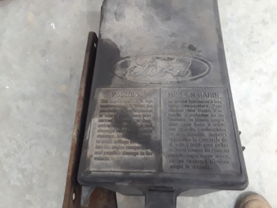 Black Ford toolbox with warning labels in English and French, mounted on a metal bracket.