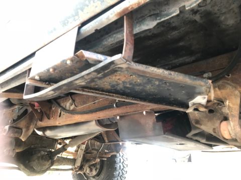 1979 Ford Bronco - The underside of a vehicle shows a rusted metal step bolted to the frame, with suspension components and exhaust visible below.