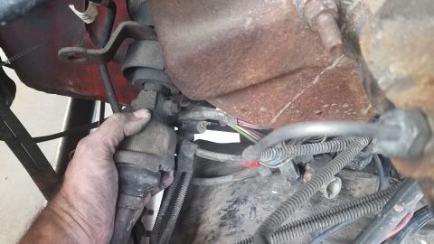 1985 Ford Bronco - A hand is shown gripping a metal component connected to a vehicle's engine bay. Wires and hoses are visible around the engine components.
