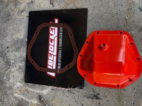 1992 Jeep Wrangler - A red differential cover and a gasket are laid out on a concrete surface next to a black booklet with the LubeLocker logo. The gasket has a…