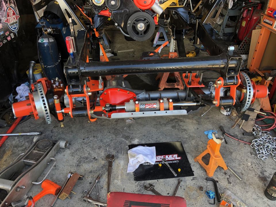 Custom off-road front axle assembly with orange suspension and disc brakes on concrete floor with tools.