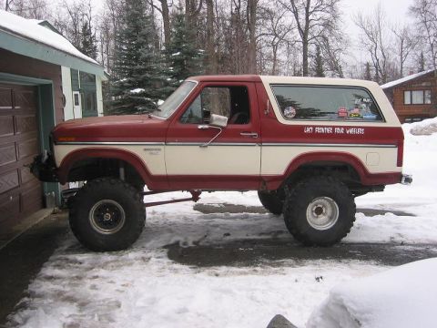 1985 Ford Bronco - This lifted Ford Bronco features a two-tone paint scheme and off-road tires, parked in a snowy environment.