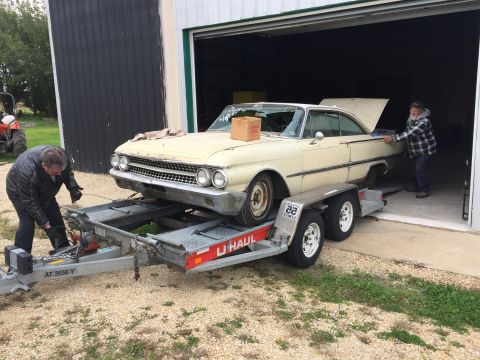 1961 Ford Starliner - A cream-colored coupe, likely from the early 1960s, is being loaded onto a U-Haul trailer in front of a garage.