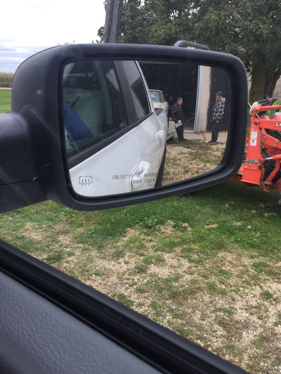 Reflection in side mirror shows two people standing near a barn and an orange tractor