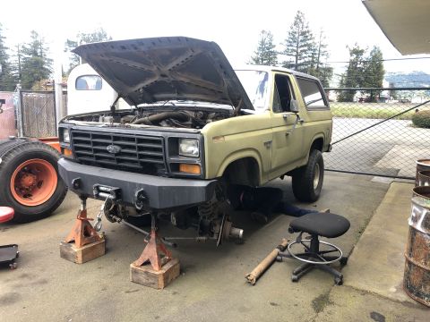 1986 Ford Bronco - Mechanic works on the front end of a 1986 Ford Bronco lifted on jack stands.