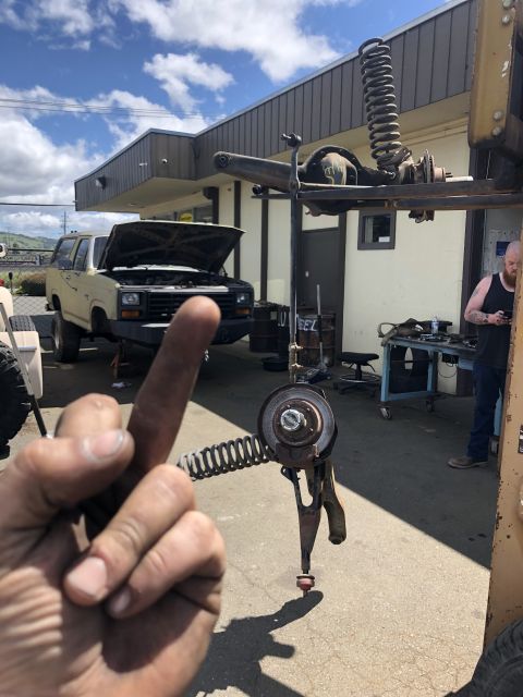 1986 Ford Bronco - A person's hand and detached suspension parts are in the foreground of a mechanic's bay with a Bronco being worked on.