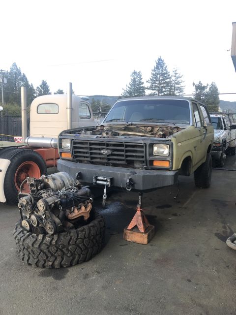 1986 Ford Bronco - SUV with its engine removed and placed on a tire, parked in front of a semi-truck.