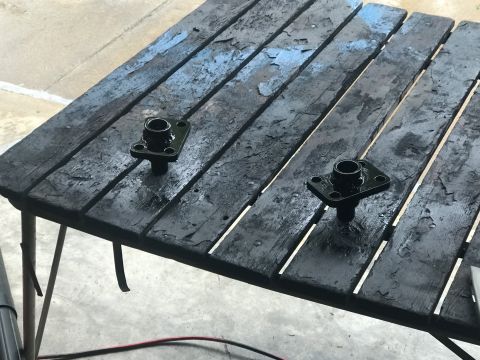 1989 Ford Bronco - Two black metal parts are displayed on a rustic, weathered wooden table, with the Supermotors logo visible at the bottom right.